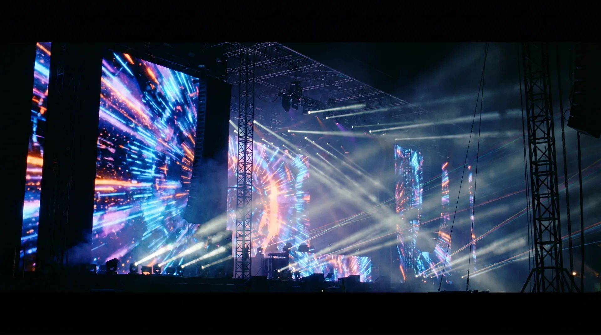 A massive concert stage at night with a blazing LED wall and light beams cutting through haze