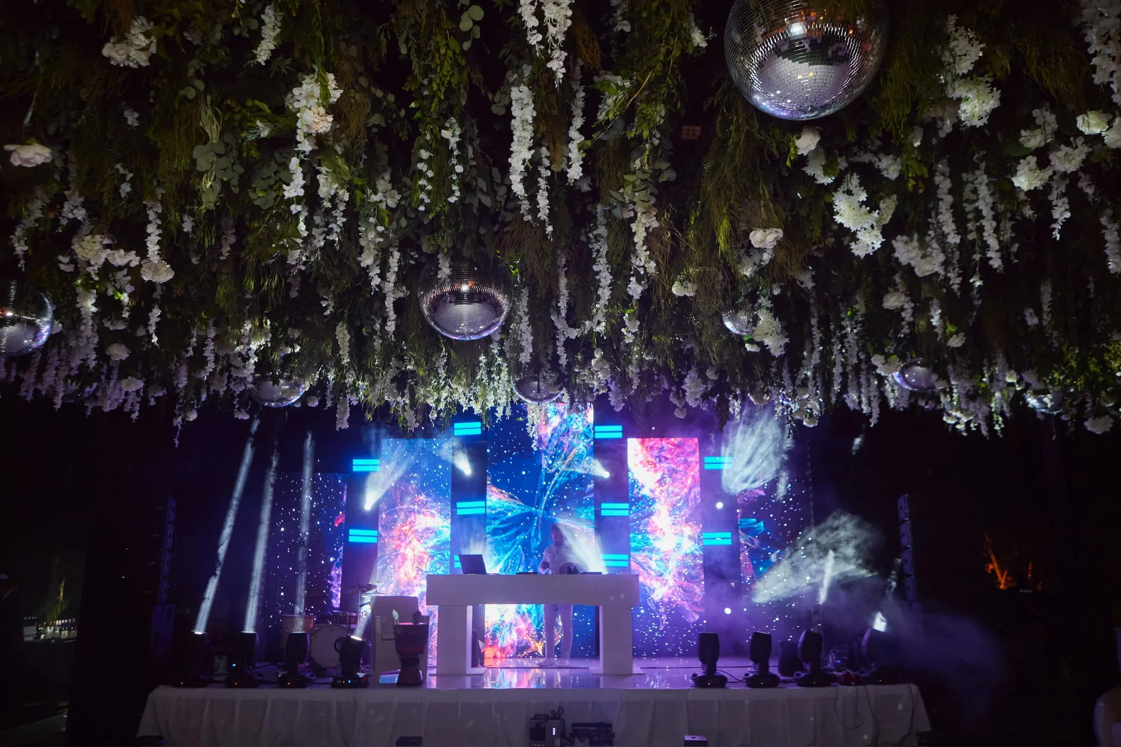 Indoor reception beneath a dense floral and greenery ceiling hung with mirror balls — a DJ booth in front of a multi-panel LED wall running abstract colour content.
