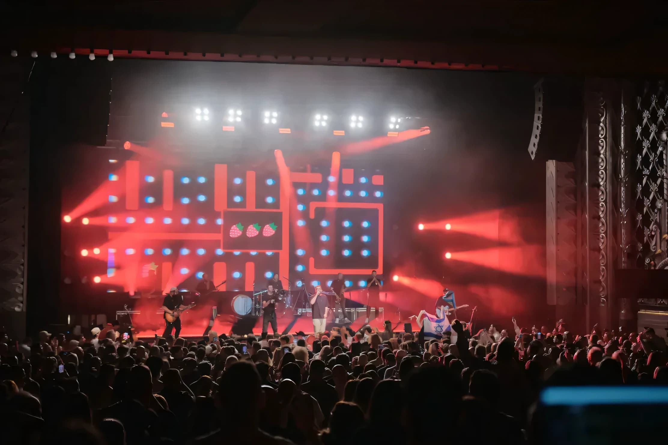 A band performing on a concert stage in red light in front of a packed crowd
