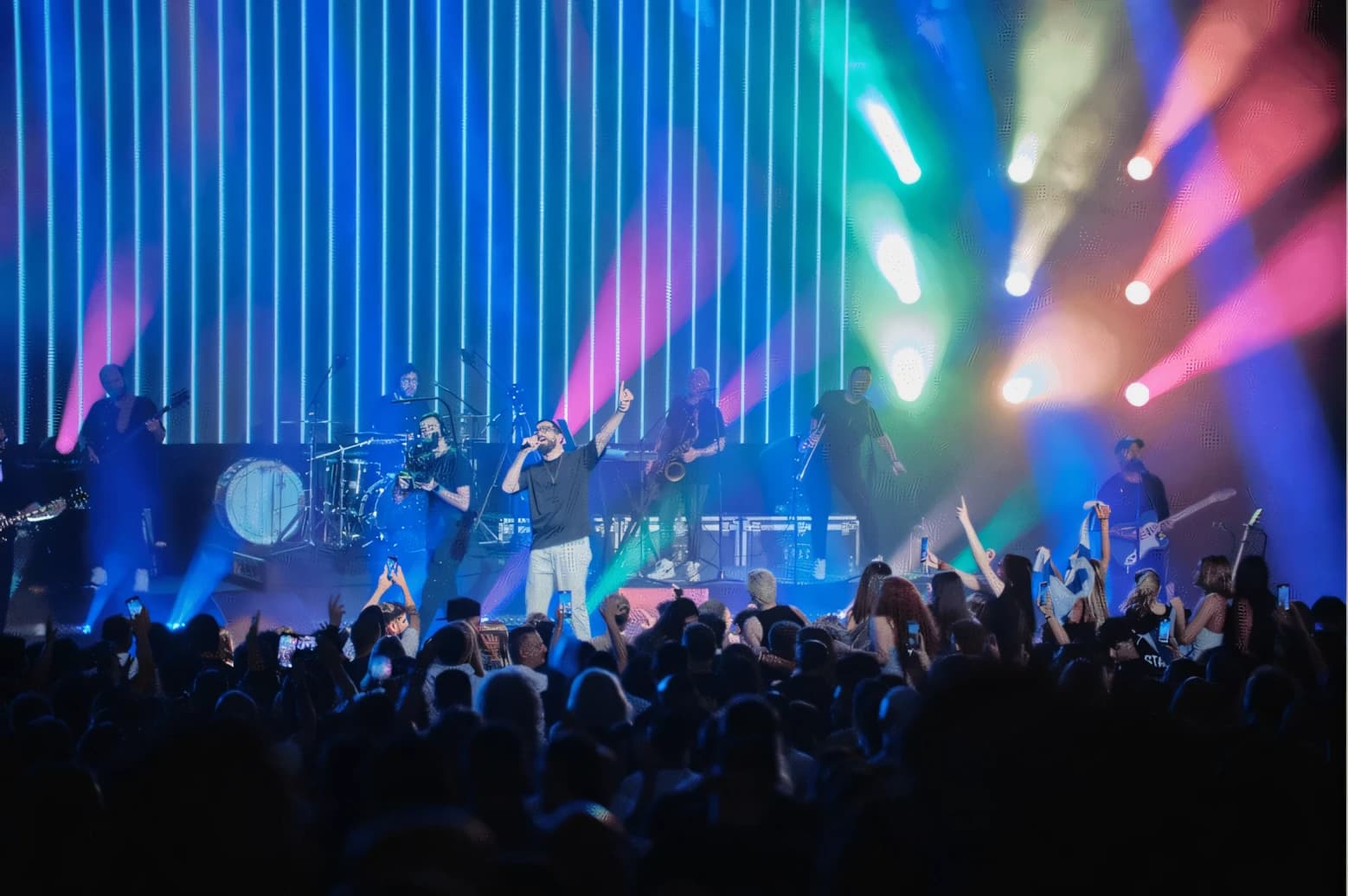Concert stage backed by a wall of vertical LED battens in blue, with a full band including saxophone and guitars, coloured beams over the crowd.