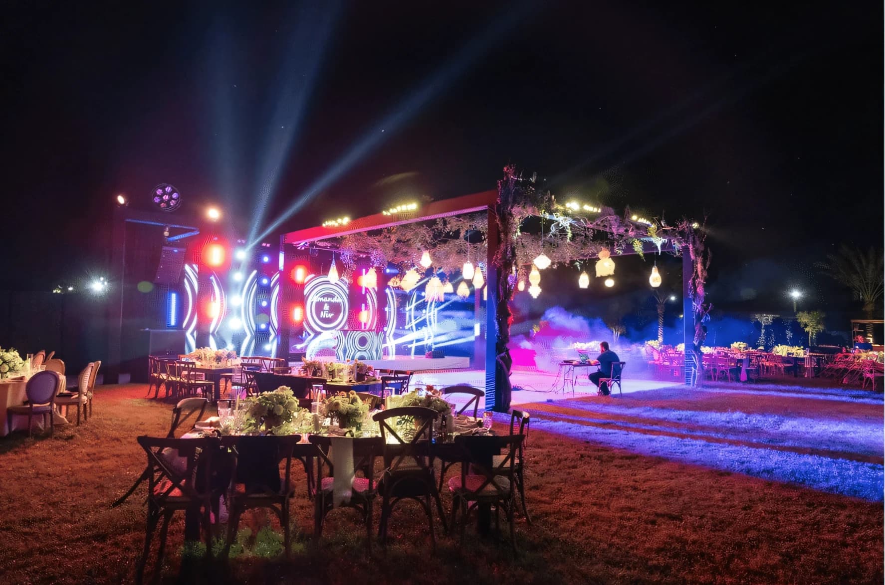 Outdoor evening private event on a lawn — a custom monogram on the LED wall, a floral arch hung with lanterns, beam lighting through haze and set dinner tables.